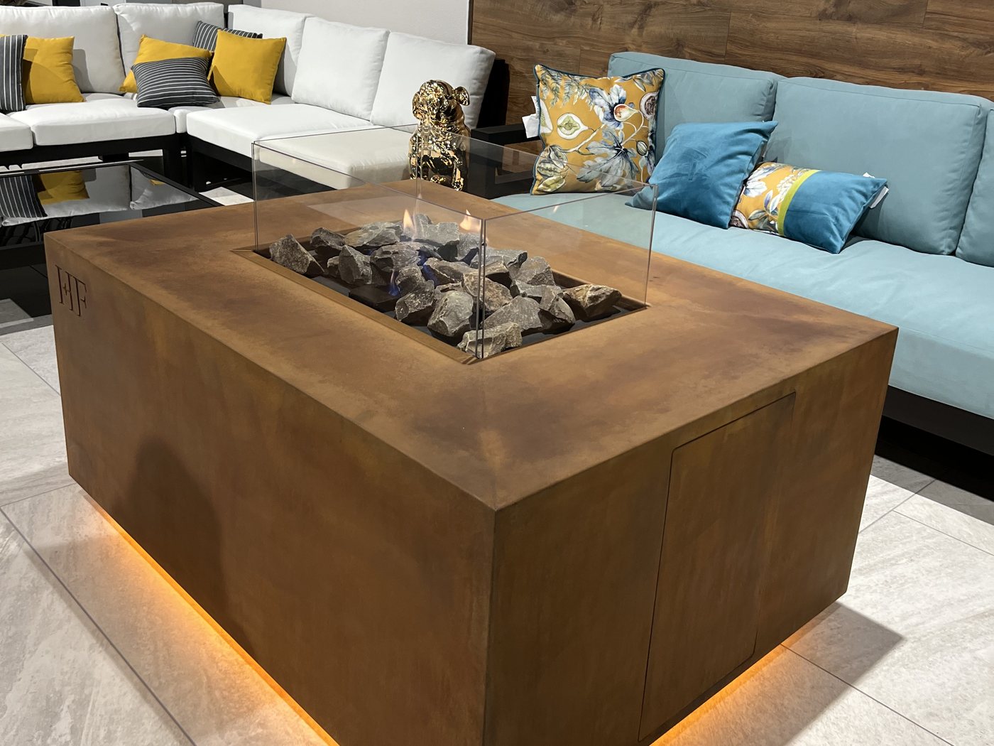 AGORA fire table in Corten steel in a lounge setting, the HF monogram embossed on the body and underlight glowing beneath