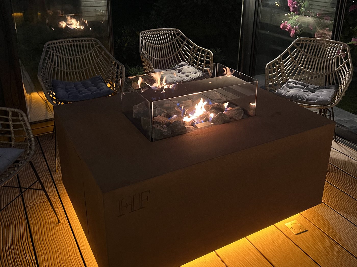 AGORA fire table in Corten steel burning on a covered terrace after dark, wicker chairs drawn up around it