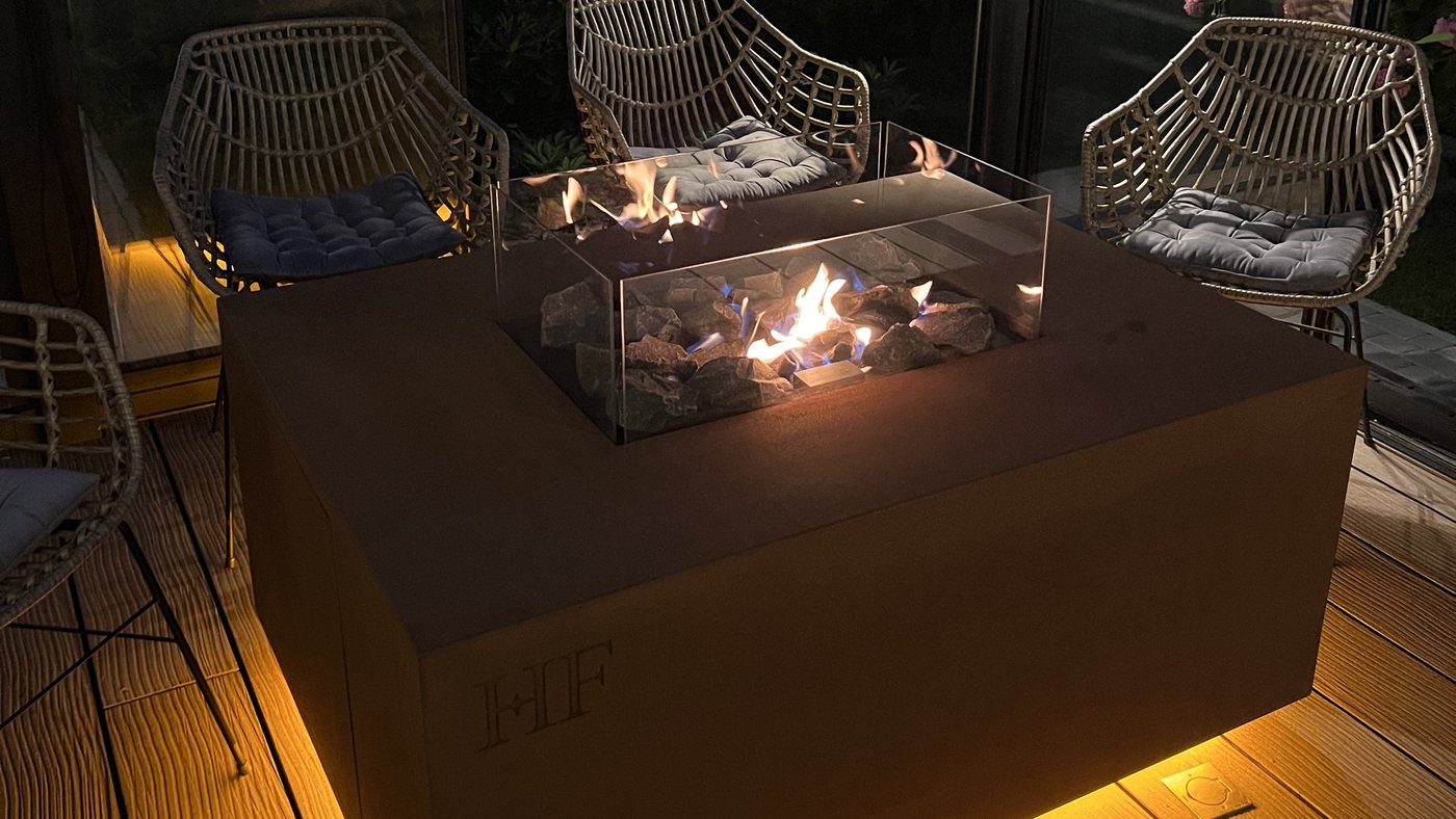 An AGORA fire table alight on a terrace at night, the flame low behind its glass shield and warm light spilling beneath the Corten body