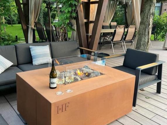 An AGORA fire table in Corten steel beneath a vine-covered pergola in summer, wine and glasses set out on the top